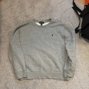 Grå sweatshirt från Polo Ralph Lauren - Klassisk grå sweatshirt från Polo Ralph Lauren med rund halsringning och broderad logga på bröstet. Tröjan har en enkel och stilren design, perfekt för en avslappnad look. Mjuk bomullskvalitet och långa ärmar.