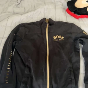 Svart jacka från Hugo Boss med gulddetaljer - Säljer en svart jacka från Hugo Boss med guldfärgad dragkedja och logga på bröstet. Jackan har lång ärm och ståkrage, samt dekorativa gulddetaljer längs ärmarna. Perfekt för dig som gillar stilrena och exklusiva plagg.