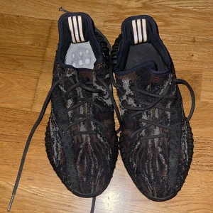 Adidas Yeezy Boost 350 V2  - Säljer ett par Adidas Yeezy Boost 350 V2 med svart och grått mönster. Skorna har snörning, rund tå och en platt sula. Ikoniska ränder på hälen och stickad ovandel för en modern look.