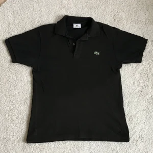 Svart pikétröja från Lacoste - Klassisk svart pikétröja från Lacoste med krage och knappar framtill. Ikoniska gröna krokodilloggan broderad på bröstet. Tillverkad i 100% bomull för en skön och luftig känsla. Perfekt för en stilren och avslappnad look. Kan gå ner i pris vid snabb affär!🔥