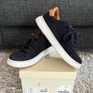 Blå mocka sneakers från Strömberg - Snygga blå mocka sneakers i nästintill nyskick från Strömberg med vita sulor och bruna detaljer vid häl och plös. Skorna har snörning och är tillverkade i mocka för en stilren look. Perfekta för skolavslutningen och om man vill ha lite extra stil i vardagen. Nypris 1 500 kr