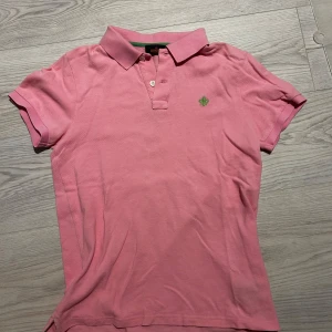 Rosa pikétröja från Morris - Säljer en klassisk rosa pikétröja från Morris med liten broderad logga på bröstet. Tröjan har korta ärmar, krage och knappar framtill. Perfekt för en stilren och avslappnad look. Fint använd skick.