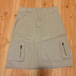 Beige kjol med fickor och dragkedjor från H&M - Säljer en beige kjol från H&M med stora fickor framtill och dragkedjedetaljer. Kjolen har en rak passform. Perfekt för en avslappnad och stilren look.