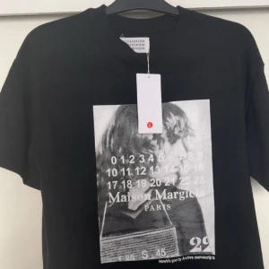  Maison Margiela t shirt - Svart t-shirt från Maison Margiela med stort grafiskt tryck framtill i svartvitt. Trycket har siffror, text och en bild på två personer. Klassisk passform och rund halsringning. Perfekt för dig som gillar statement-plagg.
