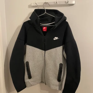 Nike hoodie jacka i svart och grå - Snygg hoodie jacka från Nike i svart och ljusgrå färg. Jackan har dragkedja framtill, två fickor med dragkedja och en vit Nike-logga på bröstet. Perfekt för en sportig och avslappnad stil. Använd 2 gånger.