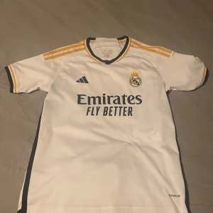 Real Madrid hemmatröja 2023/24 - Vit Real Madrid fotbollströja från Adidas med gula och svarta detaljer. Tröjan har klubbmärke och sponsortryck på bröstet samt tre gula ränder på axlarna. Klassisk V-ringning och korta ärmar.