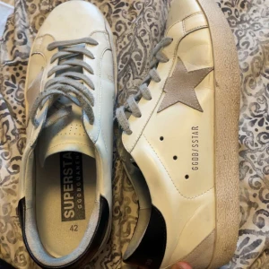 Vita sneakers från Golden Goose Superstar - Säljer ett par vita Golden Goose Superstar sneakers med grå stjärndetalj på sidan, grå snörning och svart häl. Skorna har rund tå och platt sula. Klassisk och stilren design med märkets logga på sidan. Hör av dig vid minsta lilla fråga🙌🏽