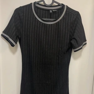 Svart ribbad klänning med vita detaljer - Svart ribbad klänning med korta ärmar och vita ränder vid halsringning och ärmslut. Klänningen har en kroppsnära passform och är perfekt för en stilren look. Enkel och snygg design som passar till många tillfällen.