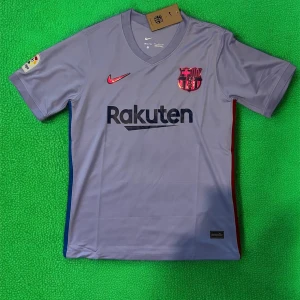 FC Barcelona bortatröja Nike - Snygg ljuslila FC Barcelona bortatröja från Nike med klubbmärke och Rakuten-tryck på bröstet. Röd Nike-logga och detaljer i blått och rött på sidorna. Kortärmad modell med V-ringning, perfekt för fotbollsfans.