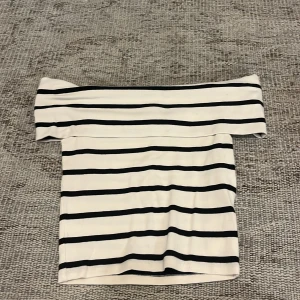Randig offshoulder topp - Säljer en vit offshoulder topp med svarta horisontella ränder. Toppen har en tight passform och är perfekt för en trendig look. Mjuk och stretchig känsla, passar snyggt till både jeans och kjol.
