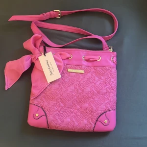 Rosa axelväska med rosett från Juicy Couture - Säljer en superfin rosa axelväska från Juicy Couture med quiltad framsida och en stor rosett på sidan. Väskan har guldfärgade detaljer, justerbar axelrem och dragkedja upptill. Perfekt för dig som vill sticka ut med en färgglad accessoar!