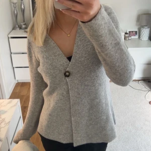 Ljusgrå kofta med knapp - Mysig ljusgrå kofta med v-ringning och en stor knapp framtill. Tröjan har lång ärm och är i ett mjukt, stickat material. Perfekt för lager-på-lager och enkel att matcha med olika outfits.