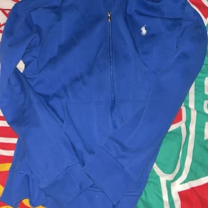 Blå hoodie från Ralph Lauren - Säljer en blå hoodie från Ralph Lauren med dragkedja framtill och vit broderad logga på bröstet. Tröjan har långa ärmar, huva och fickor framtill. Perfekt för en avslappnad stil.