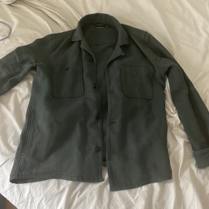 Mörkgrön overshirt från Dressmann - Säljer en mörkgrön overshirt från Dressmann i storlek S. Skjortan har två stora bröstfickor med lock, knäppning framtill och klassisk krage. Perfekt att bära som lager över en t-shirt. Materialet känns slitstarkt och har en lite grövre struktur.