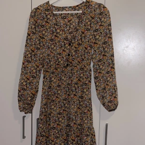 Blommig långärmad klänning med knappar - Säljer en långärmad klänning med blommigt mönster. Klänningen har v-ringning och knappar framtill, samt markerad midja för en feminin siluett. 