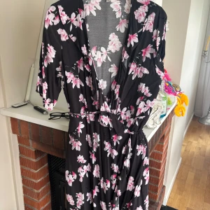 Svart blommig omlottklänning med bälte - Säljer en svart omlottklänning med rosa och vita blommor. Klänningen har v-ringning, knytband i midjan och halvlånga ärmar. Perfekt för vår och sommar när du vill känna dig både bekväm och snygg.
