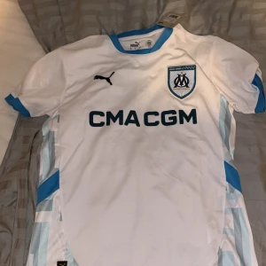 Olympique de Marseille fotbollströja - Säljer en vit kortärmad fotbollströja från Puma med blå detaljer och Olympique de Marseille-logga på bröstet. Tröjan har sponsortryck 'CMA CGM' och blå inslag längs sidorna samt runt kragen. Helt ny aldrig använd