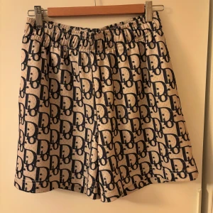 Beige och marinblå shorts från Bravest Studios - Snygga shorts från Bravest Studios i beige med marinblått mönster över hela tyget. De har elastisk midja och avslappnad passform, perfekta för varma dagar. Stora logotypmönster ger en trendig look.