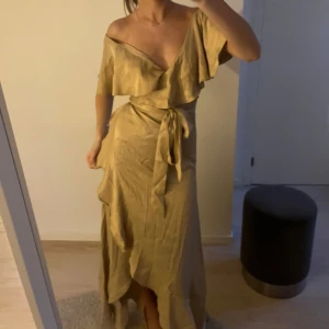 Champagnefärgad omlottklänning med volang - Säljer en champagnefärgad omlottklänning med volangdetaljer och knyt i midjan. Klänningen har offshoulder-modell och är tillverkad i ett glansigt material som ger en lyxig känsla. Perfekt för dig som vill sticka ut med en elegant look.