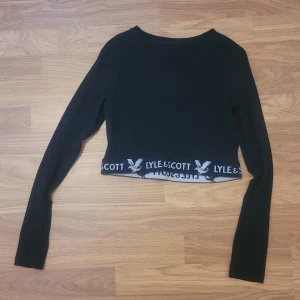 Svart croppad tröja från Lyle & Scott - Säljer en svart croppad långärmad tröja från Lyle & Scott. Tröjan har logga och text längs den nedre kanten och är perfekt för en avslappnad stil. Mjuk och skön med tight passform.