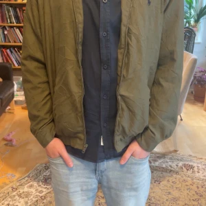 Olivgrön bomberjacka från Polo Ralph Lauren - Säljer en olivgrön bomberjacka från Polo Ralph Lauren. Jackan har dragkedja, klassisk krage och resår vid midja och ärmslut. Insidan har rutigt foder i blått och vitt. Perfekt för dig som gillar stilren och tidlös design.