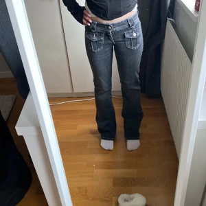 Svarta bootcut jeans med fickdetaljer - Säljer ett par svarta bootcut jeans med snygga fickor framtill och dekorativa knappar. Jeansen har låg midja och kontrastsömmar som ger en cool look. Perfekta för dig som gillar en avslappnad men trendig stil.