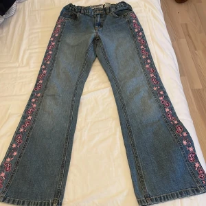 Blå jeans med broderade blommor från Old Navy i storlek 12år - Säljer ett par blå jeans från Old Navy med broderade rosa och lila blommor längs sidorna. Byxorna har elastisk midja och klassisk femficksdesign. Perfekta för dig som vill ha något unikt och färgglatt i garderoben! Mått: 27cm tvärs över på midjan 65cm på inre benet. 