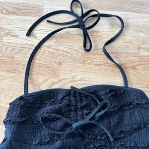 Svart volang tubtopp från SHEIN - Svart tubtopp från SHEIN med snygga volangdetaljer och knytband i nacken. Toppen har en cool struktur med vågiga rader och är axelbandslös, perfekt för att skapa en trendig look. Materialet känns lätt och luftigt, och toppen är croppad för en modern vibe.