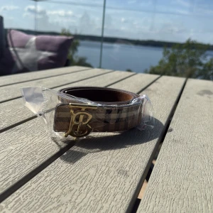 Burberry bälte - Snyggt bälte från Burberry med klassiskt rutigt mönster i beige, svart, vitt och rött. Bältet har ett stilrent guldigt spänne med Burberrys ikoniska monogram. Perfekt accessoar för att lyfta din outfit med en lyxig touch. 