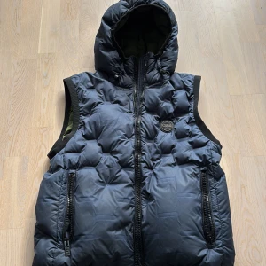 Mörkblå dunväst från Colmar - Säljer en mörkblå dunväst från Colmar Originals med huva och två stora dragkedjefickor. Västen har quiltad design och Colmar-logga på bröstet. Insidan är olivgrön och materialet är polyester med dunfyllning. Perfekt för kalla dagar och snygg till streetwear.