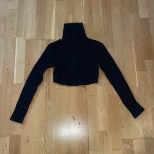 Svart croppad polotröja med snörning - Svart croppad polotröja med lång ärm och hög krage. Tröjan har ribbad struktur och snörning baktill som ger en cool detalj. Perfekt för dig som gillar trendiga och bekväma plagg med lite edge.