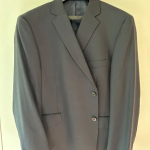 Svart kavaj från Dressmann i ull - Klassisk mörkblå kavaj från Dressmann i Super 100's ull. Tvåknäppt framtill, med tre fickor och fyra svarta knappar vid ärmslutet. Snyggt skräddad med slät yta och fodrad insida. Perfekt för dig som gillar stilren och tidlös look.