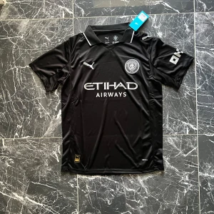 Manchester City svart fotbollströja Puma - Snygg svart Manchester City fotbollströja från Puma med vit logga och detaljer. Tröjan har klassisk krage med vit kant, korta ärmar och tryckta sponsorloggor på bröstet och ärmen. Tillverkad i lätt, ventilerande polyester som passar perfekt på planen.