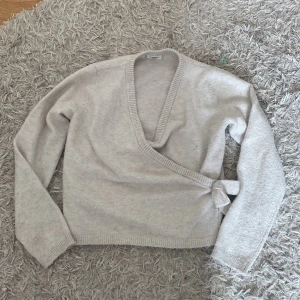 Beige omlott kofta från Kappahl - Säljer en mjuk och mysig beige omlott kofta från Kappahl med lång ärm och ribbade muddar. Koftan har v-ringad hals och knyt i sidan för en snygg passform. Perfekt att slänga på sig när det är lite kyligt ute.💕