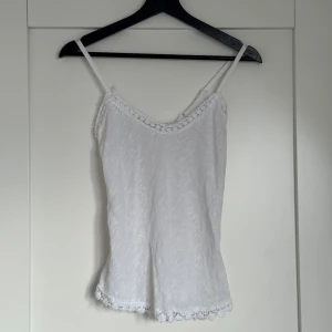 Vit linnetopp med spetsdetaljer - Supersöt vit linnetopp med smala axelband och spetsdetaljer längs halsringning och nederkant. Toppen har en lätt, luftig känsla och är perfekt för varma dagar. Spetsen ger en romantisk vibe och toppen är ärmlös för en chill look.