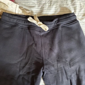 RUSSEMERCH RUSSEDRESS - Säljer ett par svarta mjukisbyxor med vit snörning i midjan. Byxorna har en relaxed passform och är gjorda i mjukt bomullsmaterial, perfekta för chill dagar eller häng med kompisar. Midjan är elastisk och byxorna har raka ben.