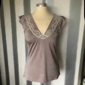 blus med spetsdetalj - Supersöt blus med v-ringning och broderad spets runt halsen. Ärmlös modell med små volanger vid axlarna som ger en gullig touch. Mjukt material som känns skönt mot huden och passar perfekt till jeans eller kjol.