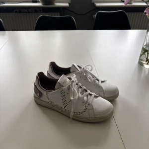 Valentino backnet vita sneakers - Säljer ett par snygga vita Valentino backnet sneakers med coola nitar på hälen och perforerat V-mönster på sidan. Skorna har snörning, rund tå och beige detaljer. Tillverkade i skinn med mjuk mesh på insidan för extra komfort. Perfekt för dig som gillar exklusiv streetstyle.