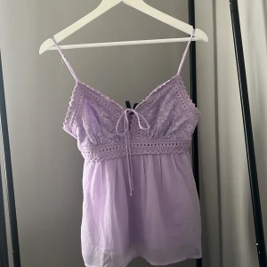 Ljuslila topp med spetsdetaljer - Supersöt ljuslila topp från Vero Moda med tunna axelband och spets upptill. Toppen har snörning framtill, broderade detaljer och en luftig kjol i bomull. Perfekt för varma dagar och ger en romantisk vibe med sin v-ringade hals och omlottkänsla.