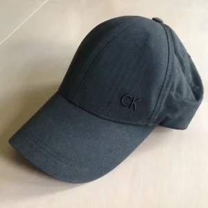 Svart keps från Calvin Klein - Snygg svart keps från Calvin Klein med diskret CK-brodyr framtill. Klassisk böjd skärm och justerbar rem med metallspänne bak. Tillverkad i bomull för skön passform och stilren look. Perfekt för dig som gillar minimalistisk design.