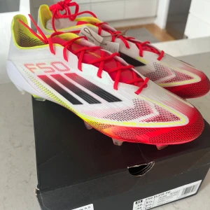 Adidas F50 Elite FG fotbollsskor - Säljer ett par Adidas F50 Elite FG fotbollsskor i en riktigt snygg färgkombo med vit bas, röda och gula detaljer samt svarta ränder. Skorna har snörning och är tillverkade i syntetmaterial för lätthet och snabbhet på planen. Perfekta för dig som vill sticka ut på matchen.