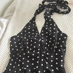 Svart prickig omlott halterneck topp - Snygg svart omlott topp med vita prickar och halterneck som knyts i nacken. Toppen har v-ringad front och en lätt volang nedtill som ger en cool siluett. Perfekt för dig som gillar retrovibbar och vill sticka ut med ett lekfullt mönster. Helt ny aldrig använd