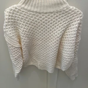 Vit polotröja med strukturstickning - Säljer en vit polotröja med grov strukturstickning och ribbad hög krage. Tröjan har långärmade ärmar och en loose passform som ger en chill vibe. Perfekt för att hålla sig varm och snygg under kyliga dagar.