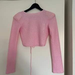 Glittrig rosa topp med snörning - Superfin bolerotopp från Gina Tricot i ljusrosa med glittrigt tyg. Croppad modell med långa ärmar och snygg snörning både i ryggen och framtill. Perfekt för att lyfta din outfit med lite extra shimmer och trendig vibe.