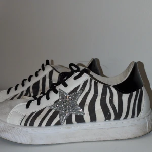 Primadonna sneakers med zebramönster - Sjukt snygga och trendiga sneakers från Primadonna Collection med svartvita zebraränder och en glittrig silverstjärna på sidan. Skorna har svarta snören, vit platt sula och svart blank detalj på hälen. Materialet är en mix av skinn och syntet, perfekt för dig som gillar att sticka ut.