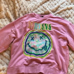 Rosa Nirvana sweatshirt med tryck - Cool rosa sweatshirt från Nirvana i storlek S. Framsidan har ett stort färgglatt tryck med bandets klassiska smiley och broderad logga i grönt och gult. Tröjan har rund hals och långa ärmar, perfekt för dig som gillar bandmerch och streetstyle.