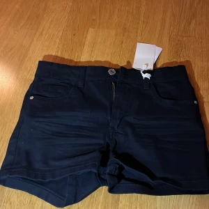 Svarta högmidjade shorts från KAPPAhl - Snygga svarta shorts från kappahl med hög midja och klassisk femficksdesign. Modellen är tight och har knapp och dragkedja framtill. Materialet är mjuk bomull med lite stretch för skön passform. Perfekta för varma dagar och enkla att matcha med din favorit-topp.