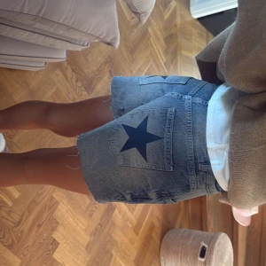 Blå jeansshorts med stjärndetalj - Snygga blå jeansshorts med råa kanter och en stor mörkblå stjärna tryckt på bakfickan. Klassisk femficksmodell med knapp och dragkedja framtill. Perfekt längd och loose fit för chill stil. Passar dig som gillar street och vill sticka ut lite extra.