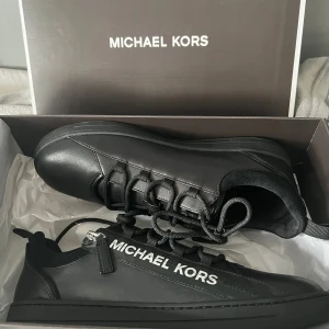Svarta sneakers från Michael Kors - Snygga svarta sneakers från Michael Kors med vit logga på sidan och MK-detalj på tungan. Skorna har rund tå, platt sula och snörning med svarta skosnören. Tillverkade i skinn med stilren design och diskreta sömmar. Perfekt för dig som gillar minimalistisk och trendig look.
