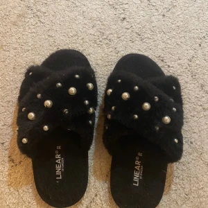 Svarta sandaler med pärlor  - Svarta sandaler från Linear of Sweden med fluffigt pälsmaterial och dekorerade med pärlor över hela ovandelen. Snyggt korslagda band ger en cool look och platt sula gör dem sköna att glida runt i. Perfekt för dig som gillar att sticka ut med detaljer.